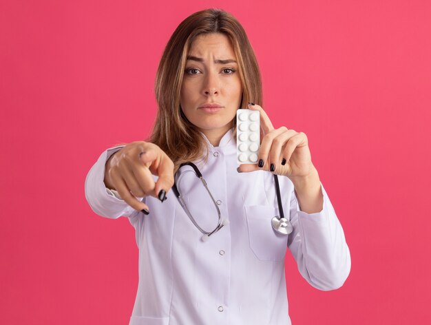 Confiant jeune femme médecin portant une robe médicale avec stéthoscope tenant des points de pilules à l'avant isolé sur un mur rose