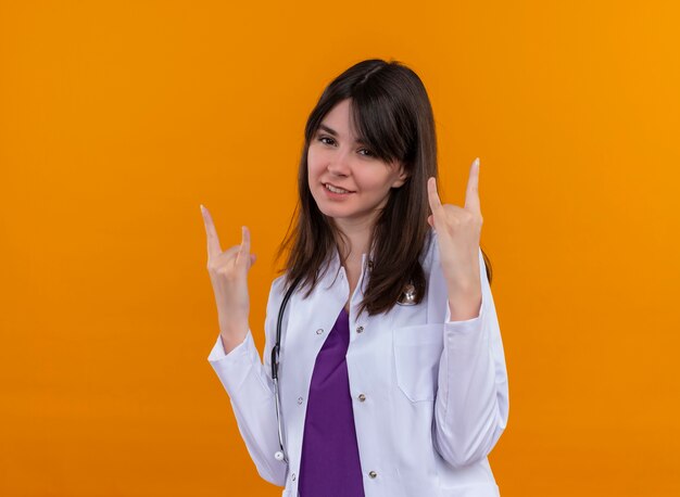 Confiant jeune femme médecin en robe médicale avec des cornes de gestes stéthoscope avec les deux mains sur fond orange isolé avec copie espace