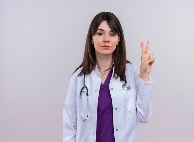 Confiant jeune femme médecin en robe médicale avec signe de victoire de gestes stéthoscope sur fond blanc isolé avec espace de copie