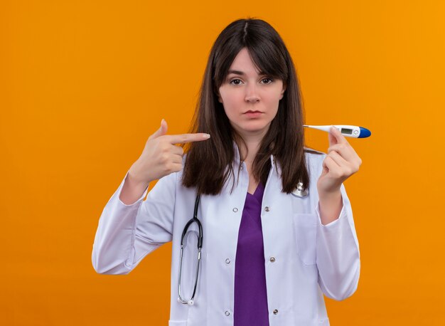 Confiant jeune femme médecin en robe médicale avec stéthoscope détient thermomètre et pointe au thermomètre sur fond orange isolé avec copie espace