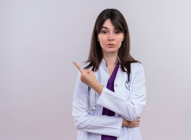 Confiant jeune femme médecin en robe médicale avec stéthoscope pointe sur le côté et regarde la caméra sur fond blanc isolé avec copie espace