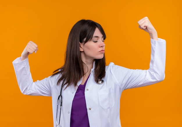 Confiant jeune femme médecin en robe médicale avec stéthoscope tend les biceps sur fond orange isolé