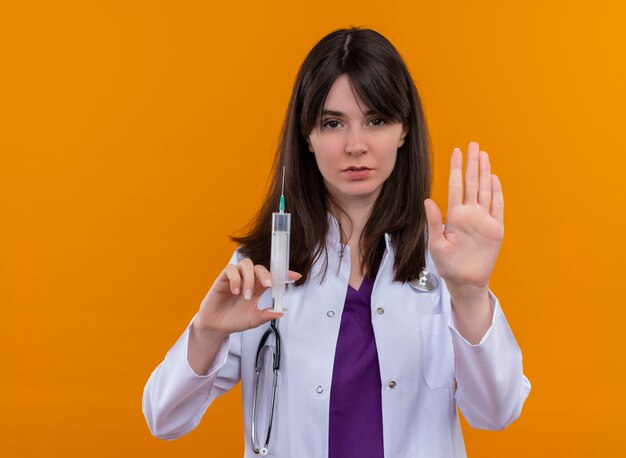 Confiant jeune femme médecin en robe médicale avec stéthoscope tient la seringue et les gestes s'arrêtent sur fond orange isolé