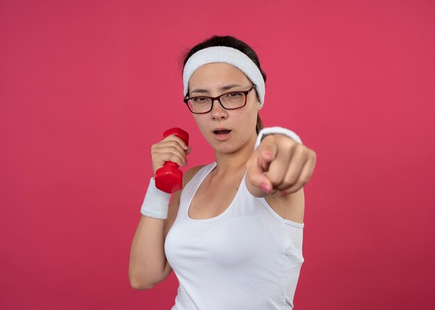 Confiant jeune femme sportive dans des lunettes optiques portant un bandeau et des bracelets tient des haltères et des points à l'avant isolé sur un mur rose