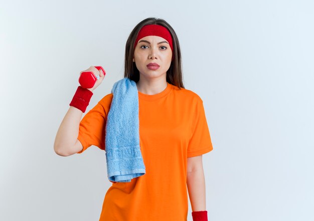 Confiant jeune femme sportive portant un bandeau et des bracelets tenant un haltère avec une serviette sur l'épaule isolé sur un mur blanc avec espace de copie