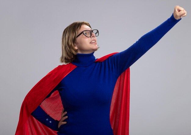 Photo gratuite confiant jeune femme de super-héros blonde en cape rouge portant des lunettes debout dans la pose de superman en regardant son poing isolé sur un mur blanc