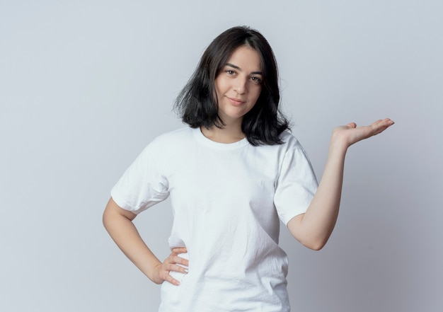 Confiant jeune fille assez caucasienne mettant la main sur la taille et montrant la main vide isolé sur fond blanc