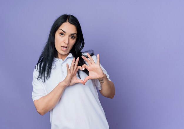 Confiant jeune fille brune caucasienne gestes signe de main coeur isolé sur mur violet