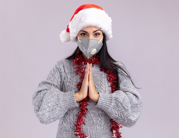 Confiant jeune fille caucasienne portant chapeau de Noël et guirlande de guirlandes autour du cou avec masque de protection regardant la caméra en gardant les mains ensemble isolé sur fond blanc