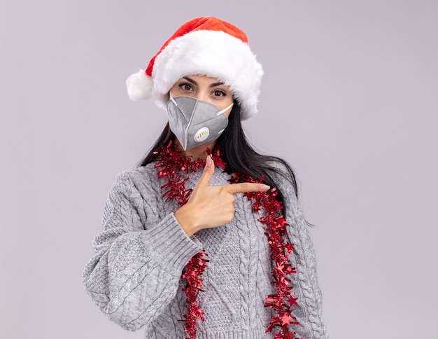 Confiant jeune fille caucasienne portant chapeau de Noël et guirlande de guirlandes autour du cou avec masque de protection regardant la caméra pointant sur le côté isolé sur fond blanc avec espace de copie