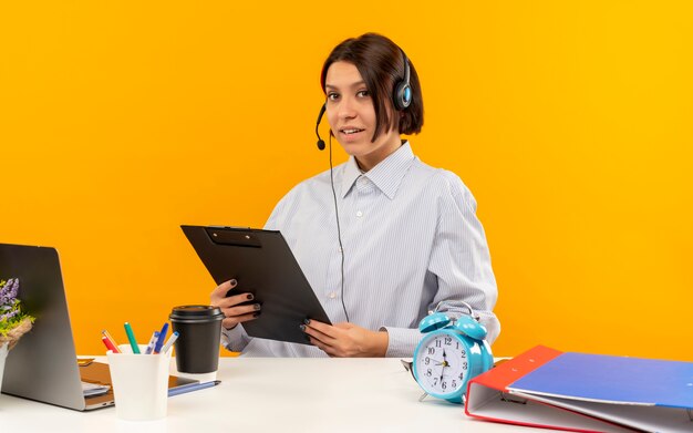 Confiant jeune fille de centre d'appels portant un casque assis au bureau tenant le presse-papiers isolé sur orange
