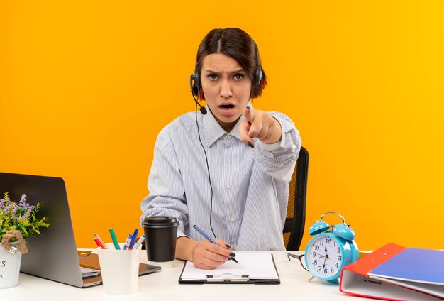 Confiant jeune fille de centre d'appels portant un casque assis au bureau tenant un stylo et pointant isolé sur orange