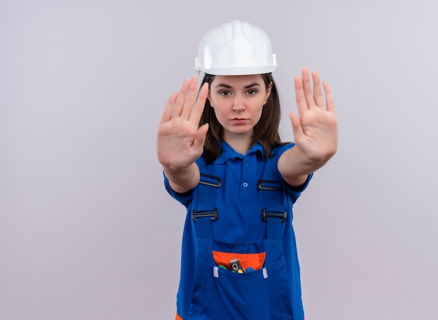 Photo gratuite confiant jeune fille constructeur avec un casque de sécurité blanc et des gestes uniformes bleus s'arrêtent à deux mains sur fond blanc isolé avec espace de copie