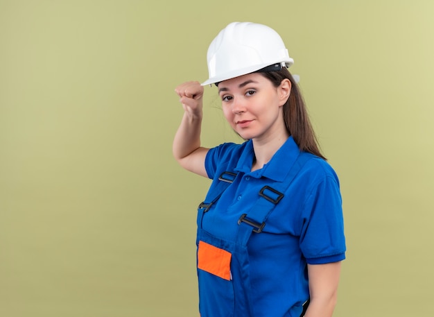 Confiant jeune fille constructeur avec casque de sécurité blanc et uniforme bleu lève le poing et regarde la caméra sur fond vert isolé avec copie espace