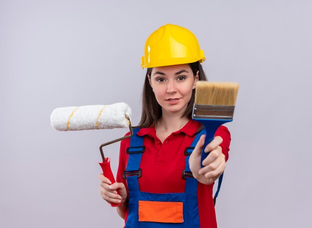 Confiant jeune fille de constructeur tient le rouleau à peinture et le pinceau sur fond blanc isolé