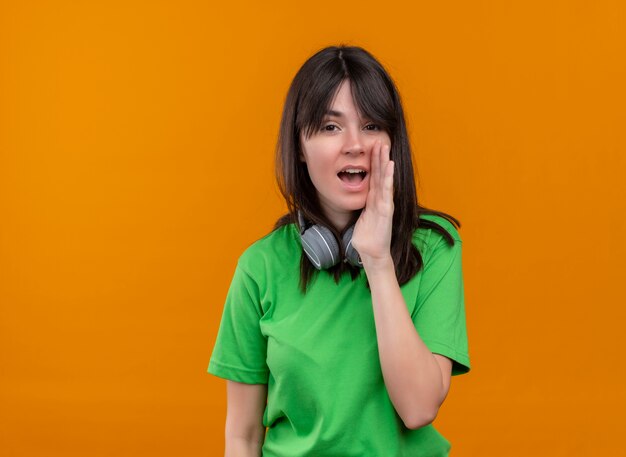 Confiant jeune fille de race blanche en chemise verte avec un casque fait semblant sur fond orange isolé