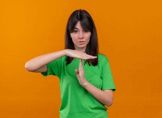 Confiant jeune fille de race blanche en chemise verte montre le geste de temps mort sur fond orange isolé