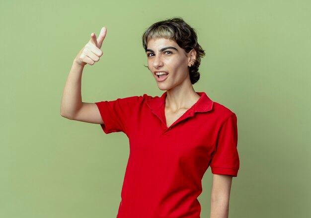 Confiant jeune fille de race blanche avec coupe de cheveux de lutin faisant le geste de pistolet avec la main et pointant vers la caméra isolée sur fond vert olive avec espace de copie