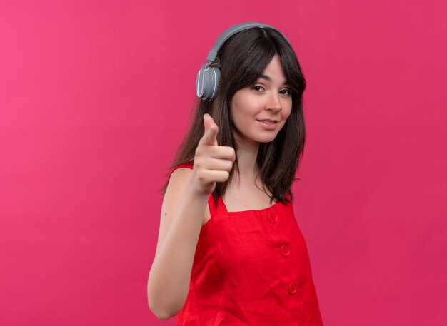 Confiant jeune fille de race blanche avec des écouteurs pointant sur la caméra sur fond rose isolé avec copie espace