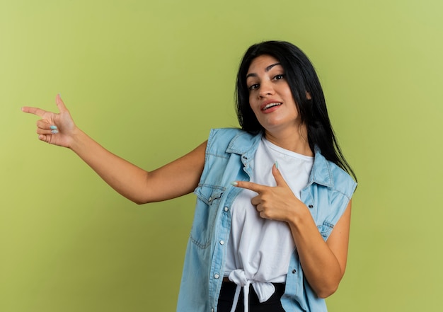 Confiant jeune fille de race blanche pointe sur le côté avec deux mains isolé sur fond vert olive avec copie espace