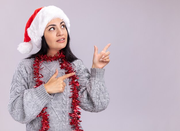 Confiant jeune fille de race blanche portant chapeau de Noël et guirlande de guirlandes autour du cou à la recherche et pointant sur le côté isolé sur fond blanc avec copie espace