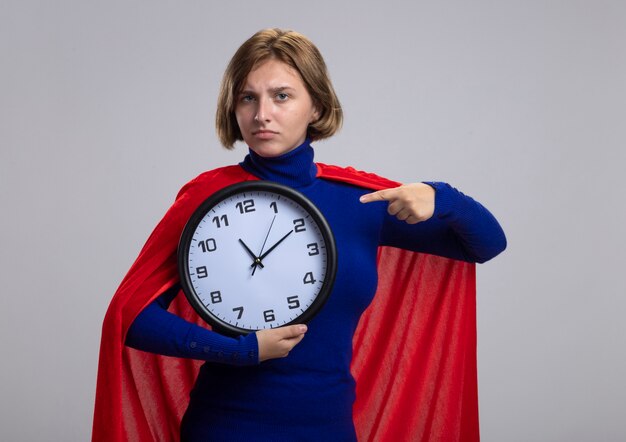 Confiant jeune fille de super-héros blonde en cape rouge tenant et pointant sur horloge isolé sur mur blanc