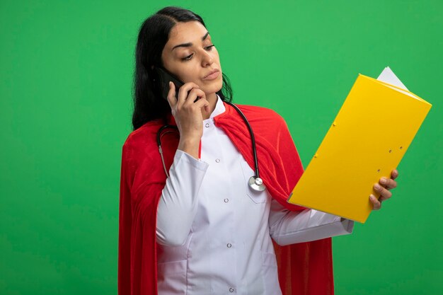 Confiant jeune fille de super-héros portant une robe médicale avec stéthoscope tenant et regardant le dossier parle au téléphone isolé sur vert