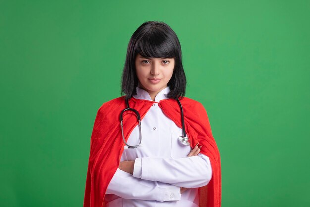 Confiant jeune fille de super-héros portant un stéthoscope avec une robe médicale et une cape croisant les mains isolées sur le vert
