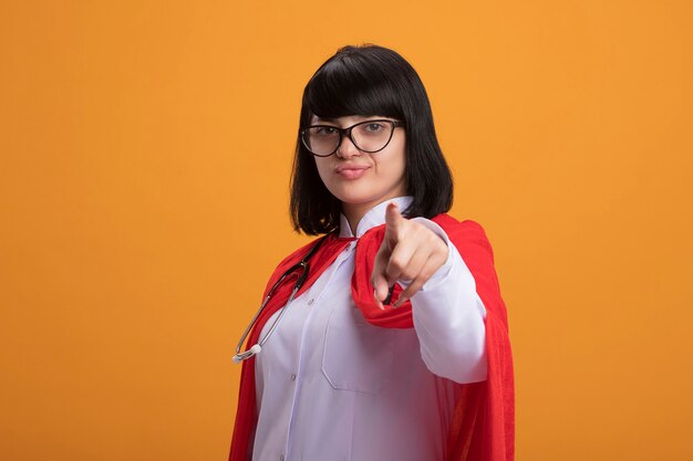 Confiant jeune fille de super-héros portant un stéthoscope avec une robe médicale et un manteau avec des lunettes vous montrant le geste isolé sur un mur orange