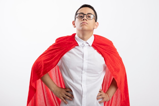 Confiant jeune garçon de super-héros en cape rouge portant des lunettes regardant la caméra en gardant les mains sur la taille isolé sur fond blanc