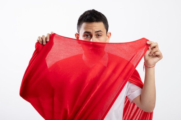 Confiant jeune garçon de super-héros en cape rouge saisissant la cape à l'avant par derrière isolé sur mur blanc