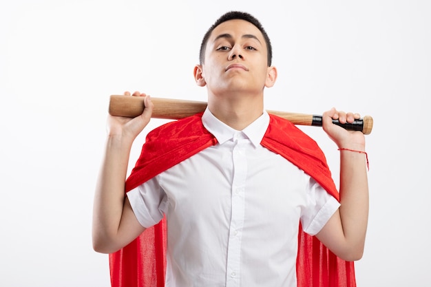 Confiant jeune garçon de super-héros en cape rouge tenant une batte de baseball derrière le cou en regardant la caméra isolée sur fond blanc
