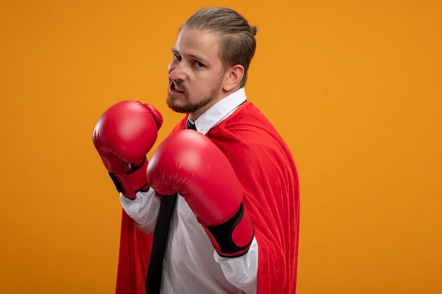 Photo gratuite confiant jeune gars de super-héros portant cravate et gants de boxe debout dans la pose de combat isolé sur fond orange avec espace de copie