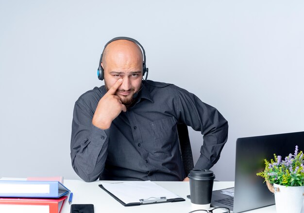 Confiant jeune homme de centre d'appels chauve portant un casque assis au bureau avec des outils de travail mettant le doigt sous les yeux isolé sur blanc