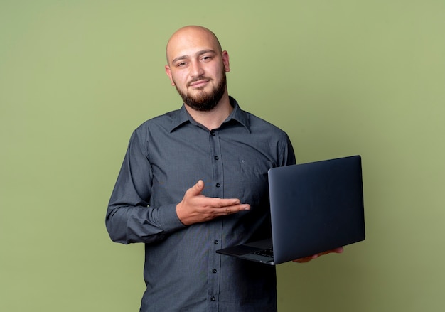 Confiant jeune homme de centre d'appels chauve tenant et pointant avec la main à l'ordinateur portable isolé sur vert olive avec espace de copie