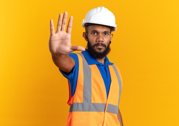 Confiant jeune homme constructeur afro-américain en uniforme avec un casque de sécurité faisant un geste d'arrêt isolé sur fond orange avec espace de copie