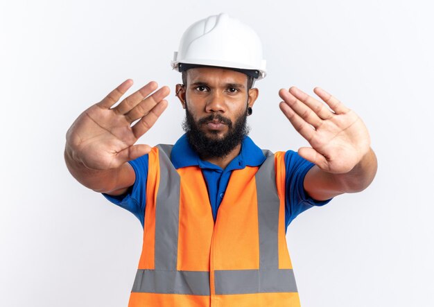 Confiant jeune homme constructeur afro-américain en uniforme avec un casque de sécurité gesticulant un panneau d'arrêt isolé sur fond blanc avec espace de copie