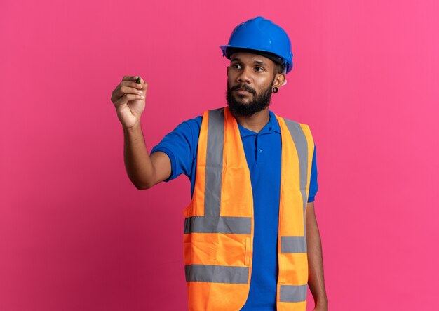 Confiant jeune homme constructeur afro-américain en uniforme avec un casque de sécurité tenant et regardant un stylo isolé sur fond rose avec espace de copie