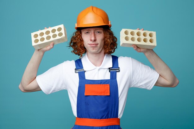 Confiant jeune homme constructeur en uniforme tenant des briques isolées sur fond bleu