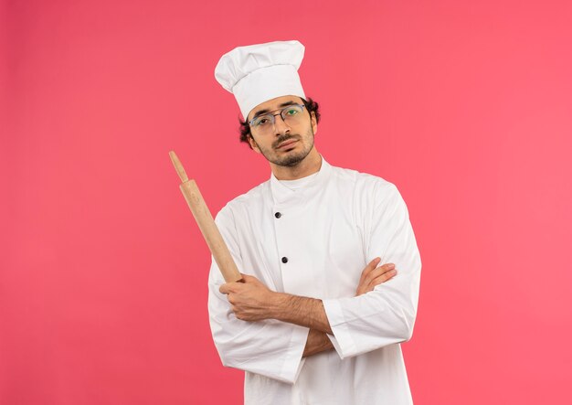 Confiant jeune homme cuisinier portant l'uniforme de chef et des lunettes croisant les mains et tenant le rouleau à pâtisserie