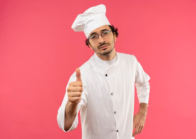 Confiant jeune homme cuisinier portant un uniforme de chef et des lunettes mettant la main sur la hanche et son pouce vers le haut