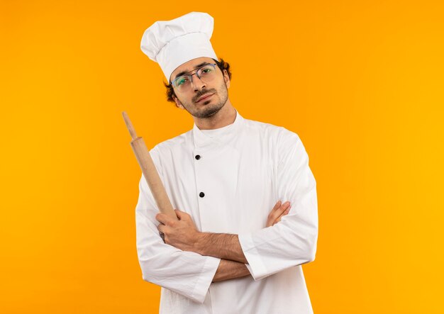 Confiant jeune homme cuisinier portant l'uniforme de chef et des lunettes tenant un rouleau à pâtisserie et croisant les mains
