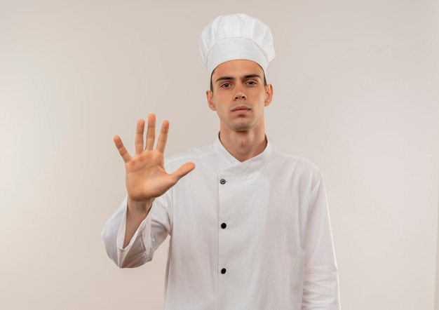 Photo gratuite confiant jeune homme cuisinier portant l'uniforme de chef montrant le geste d'arrêt sur un mur blanc isolé