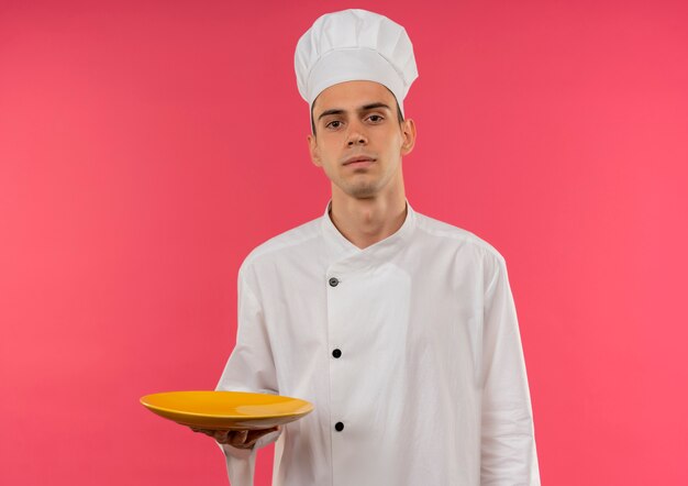 Confiant jeune homme cuisinier portant un uniforme de chef tenant la plaque sur un mur rose isolé avec copie espace