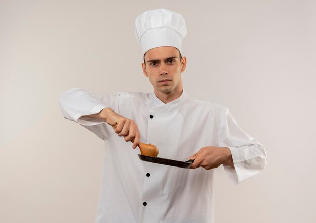 Confiant jeune homme cuisinier portant l'uniforme de chef tenant une poêle et une cuillère sur un mur blanc isolé