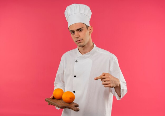 Confiant jeune homme cuisinier portant l'uniforme de chef tenant et points orange sur planche de bois sur mur rose isolé