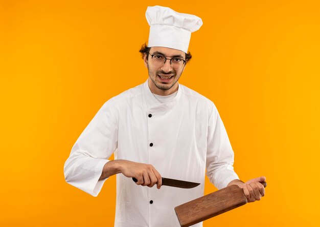 Confiant jeune homme cuisinier portant l'uniforme de chef et des verres tenant une planche à découper et un couteau