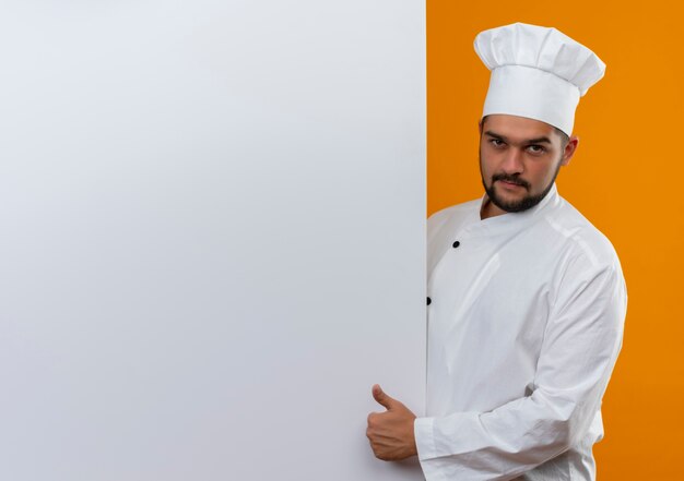 Confiant jeune homme cuisinier en uniforme de chef debout derrière un mur blanc montrant le pouce vers le haut à la recherche d'isolement sur le mur orange avec espace de copie