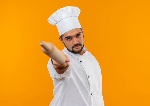 Confiant jeune homme cuisinier en uniforme de chef étirant le rouleau à pâtisserie vers isolé sur mur orange avec espace de copie