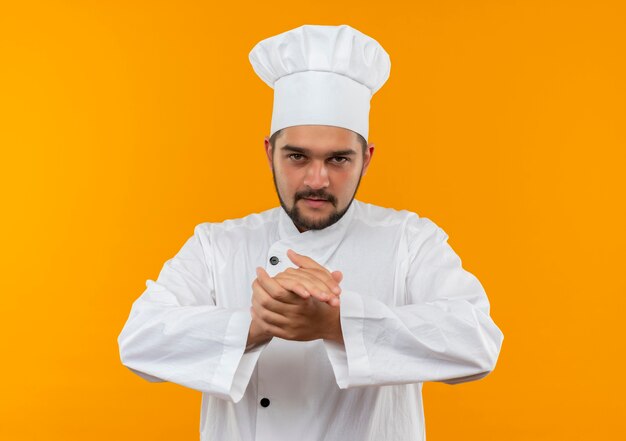 Confiant jeune homme cuisinier en uniforme de chef gardant les mains ensemble isolé sur mur orange